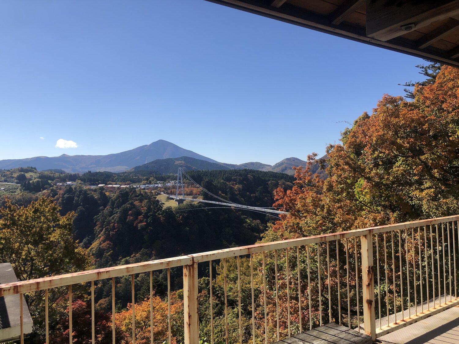 The Kokonoe Yume Otsurihashi suspension bridge seen from a distance, framed by autumn foliage with Mt. Kuju in the background